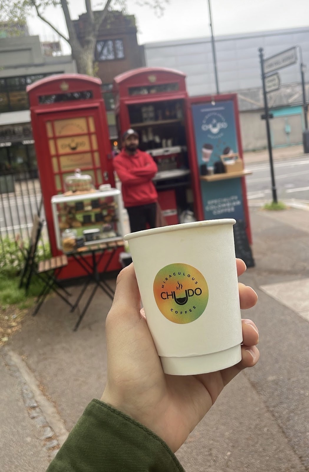 A white takeaway cup held up in front of the red phone-box kiosk